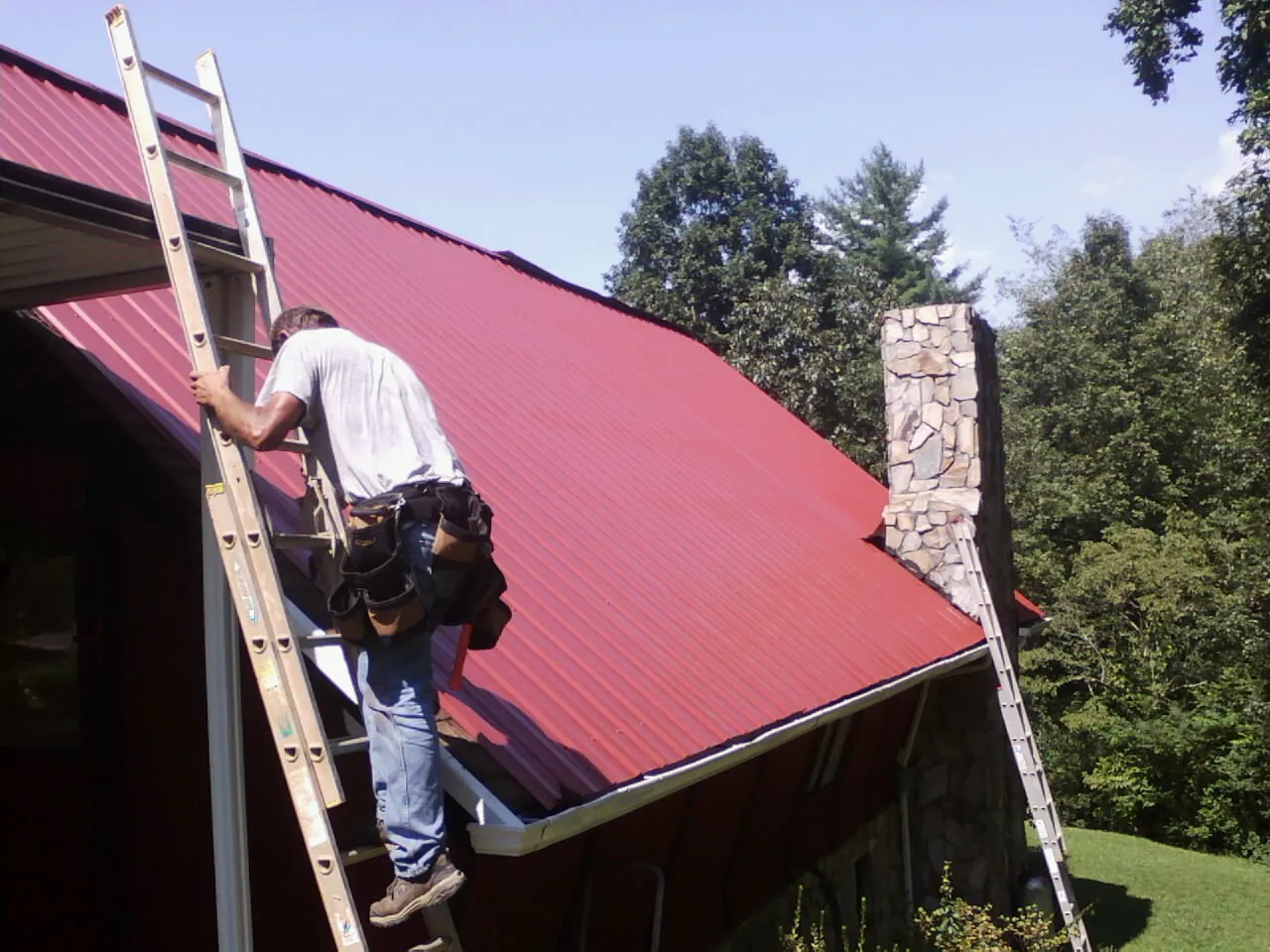 Roofer on a ladder beside a red metal roof and stone chimney — Soffit & Fascia Repair by Roofing Brothers in Howey In The Hills